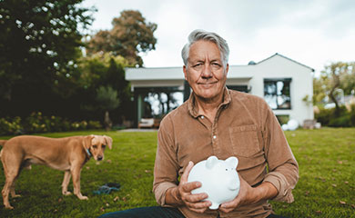 Älterer Mann sitzt im Garten und hält ein weißes Sparschwein in der Hand. Neben ihm sein Hund.